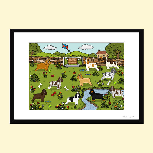 A framed art print featuring a whimsical illustration of various dog breeds in a green park with a small stream, stone wall, and a sign reading 'Welsh Corgi Welcome'. The print is presented in a black frame with a white mat against a light beige background.