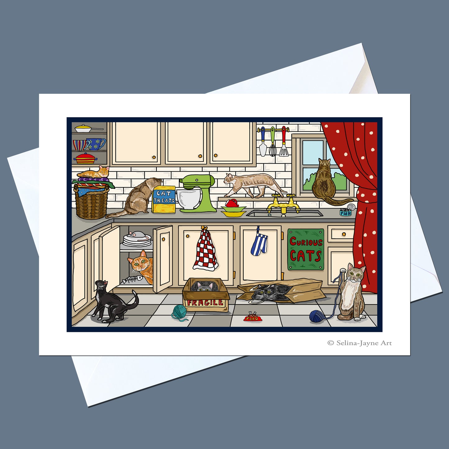 A printed art card or greeting card featuring a whimsical illustrated kitchen scene with multiple cats, shown with white envelopes on a muted blue-gray background. The artwork includes cream cabinets, a green mixer, a red curtain, and several cats in different poses.