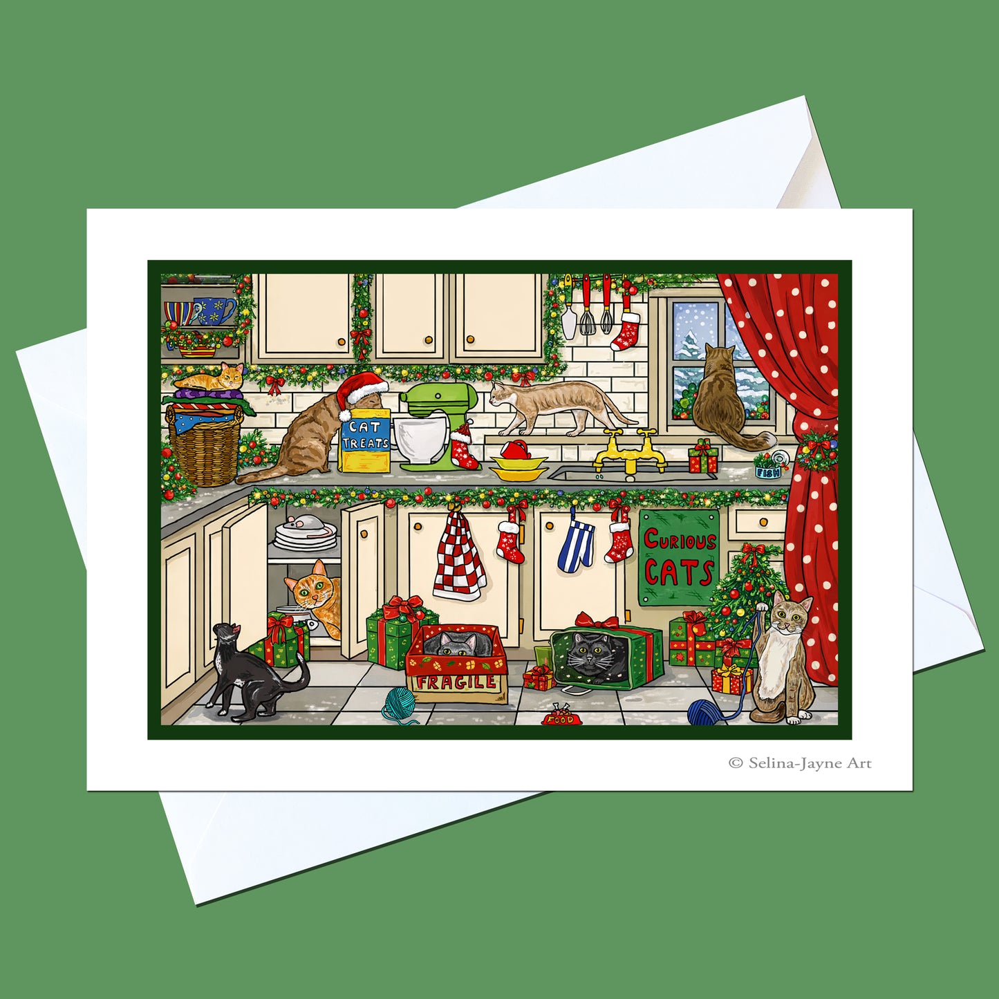A Christmas-themed kitchen art print featuring cats in a decorated kitchen, shown with a white envelope on a solid green background. The print has a white border and the artist's name 'Selina Jayne Art' is printed in the bottom right corner.
