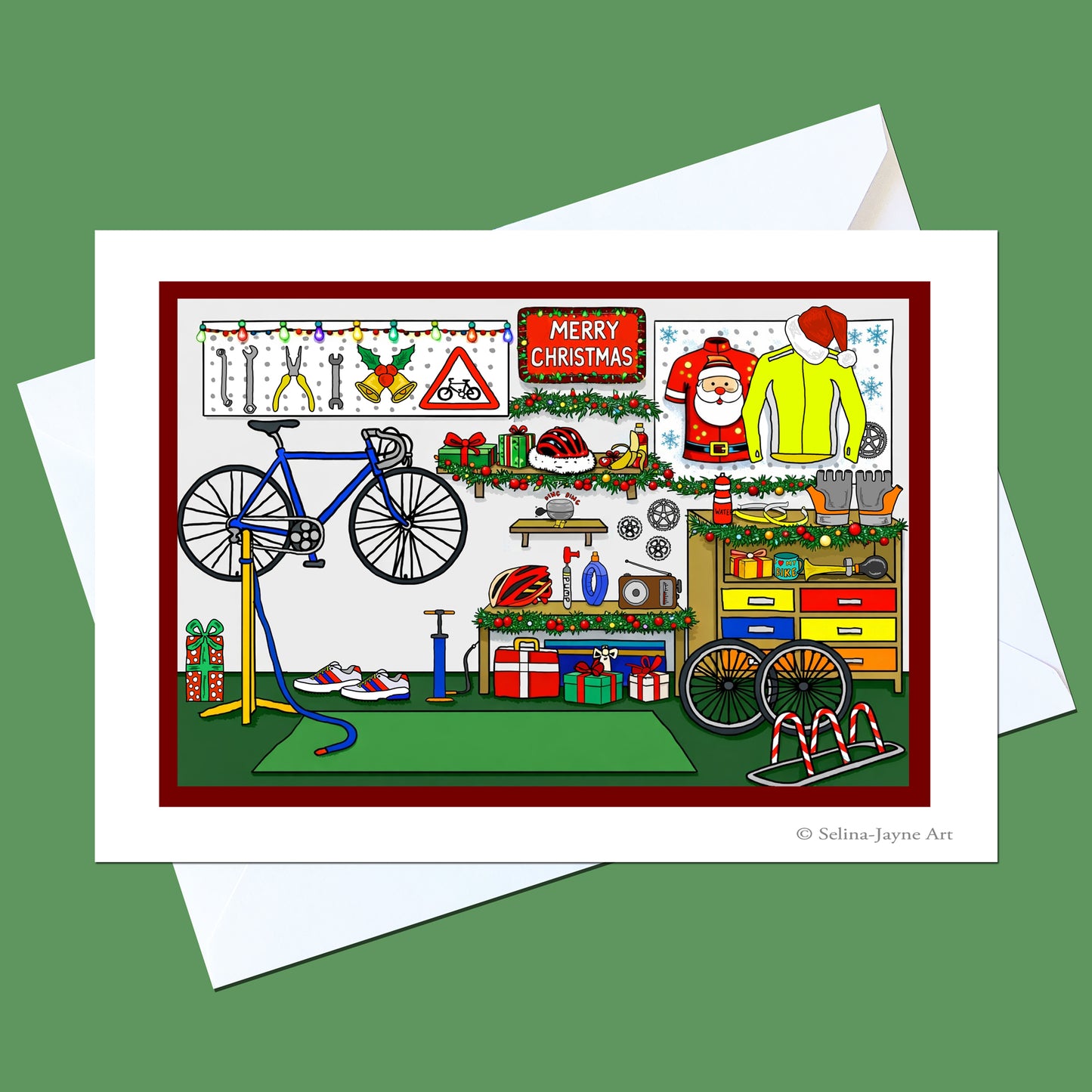 A Christmas-themed greeting card featuring a busy garage workshop scene with a blue bicycle, holiday decorations, gift boxes, and a 'Merry Christmas' sign, shown with white envelopes on a solid green background.