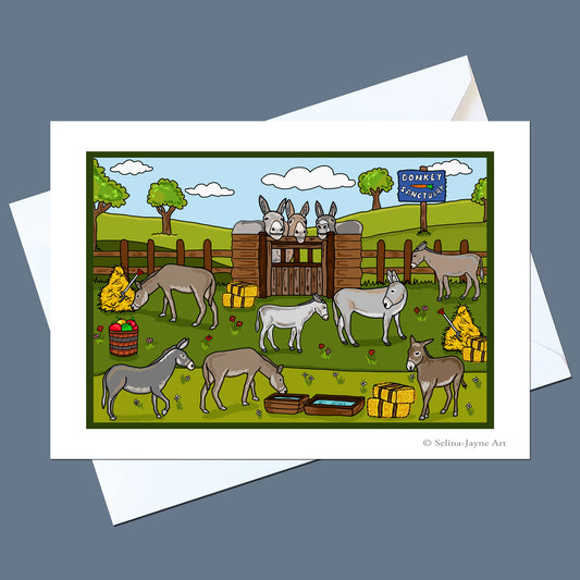A rectangular art print with a white border shows a colorful cartoon scene of donkeys in a farm paddock with a gate, fence, trees, and a blue sign. The print is displayed on top of white envelopes against a muted blue-gray background.