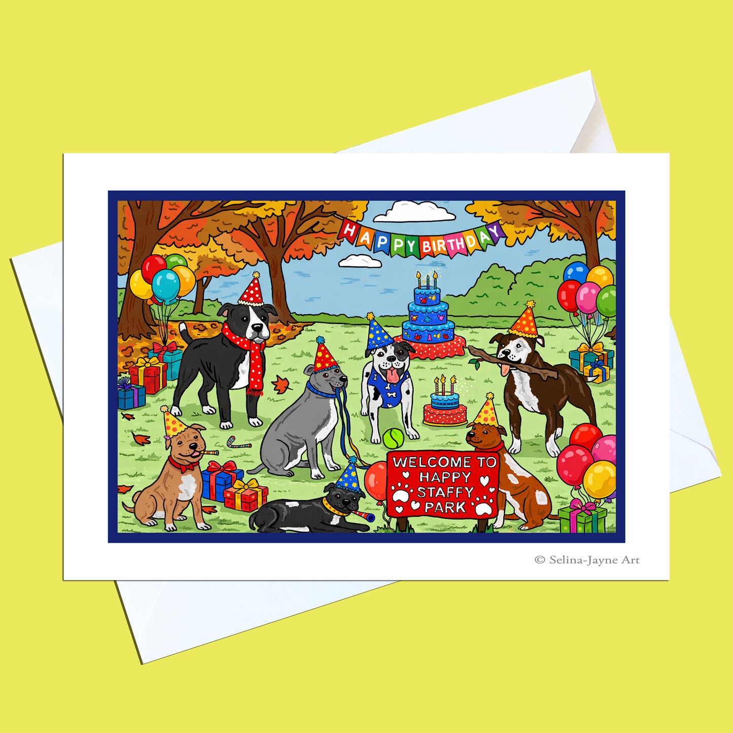 A birthday greeting card and envelope shown on a pale yellow background. The card features a colorful cartoon illustration of several dogs celebrating outdoors with a birthday cake, balloons, trees, and a sign reading 'Welcome to Happy Staffy Park'.