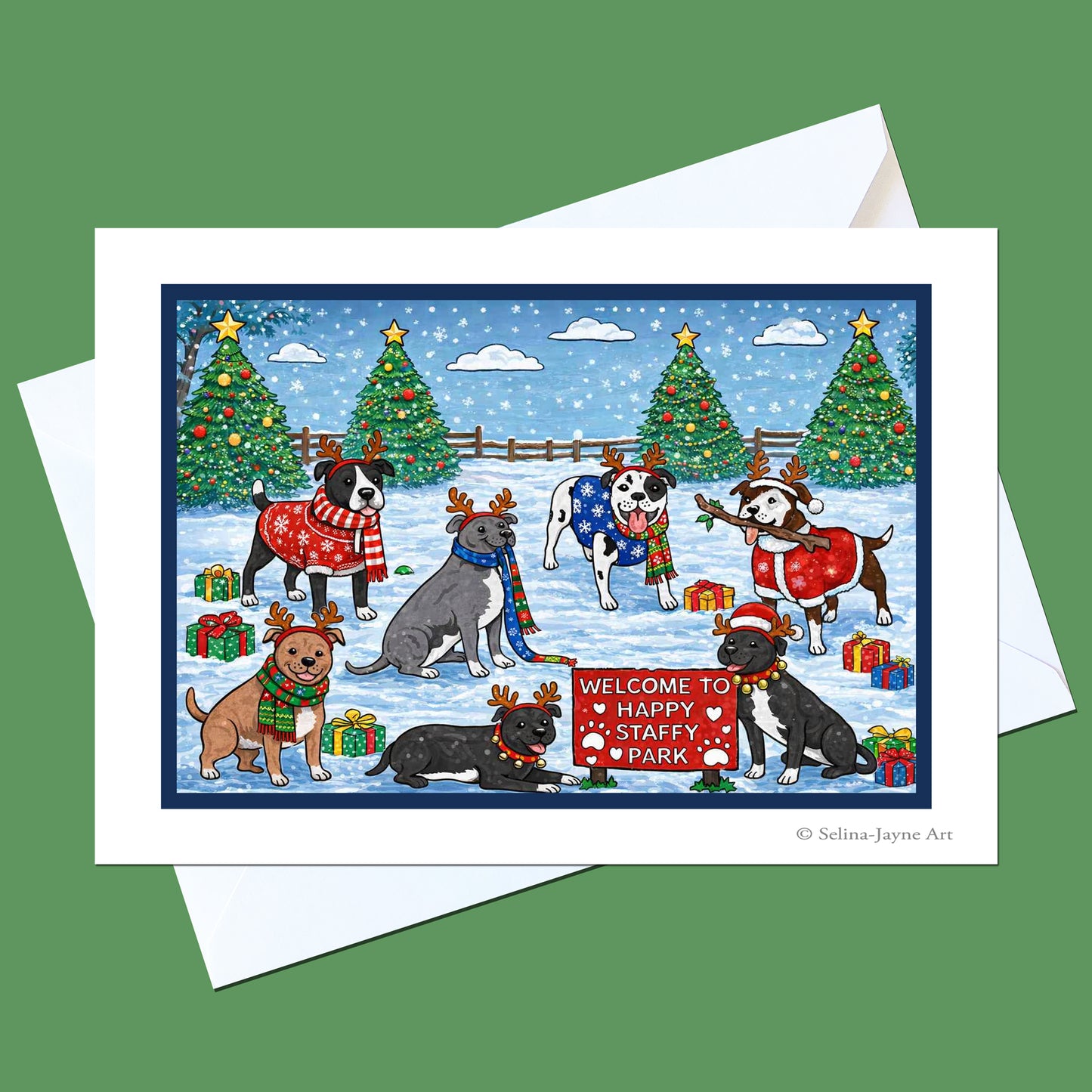 A square holiday greeting card with a white border and white envelope, shown on a solid green background. The card features a colorful illustration of several dogs dressed in Christmas sweaters and scarves, standing in a snowy winter park with decorated trees, gift boxes, and a sign that reads 'Welcome to Happy Stuffy Park'.